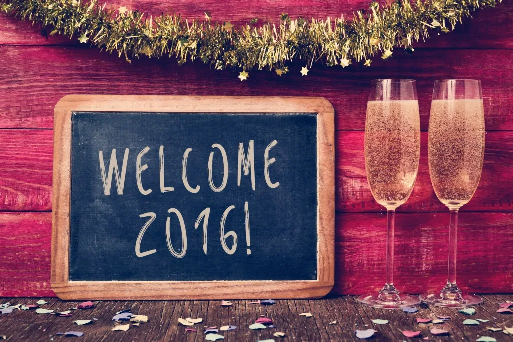 a chalkboard with the text welcome 2016 written in it, some confetti and a pair of glasses with champagne on a red rustic wooden background ornamented with tinsel